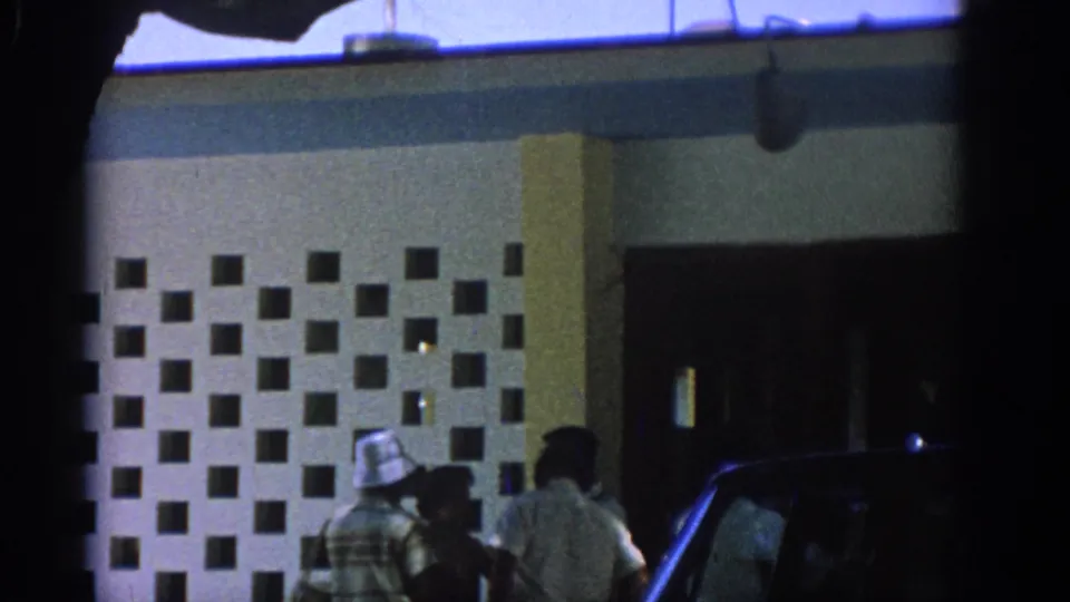 Rhodes, Greece 1962 frame with visitors near a black-and-white patterned facade and a parked dark car at the edge.