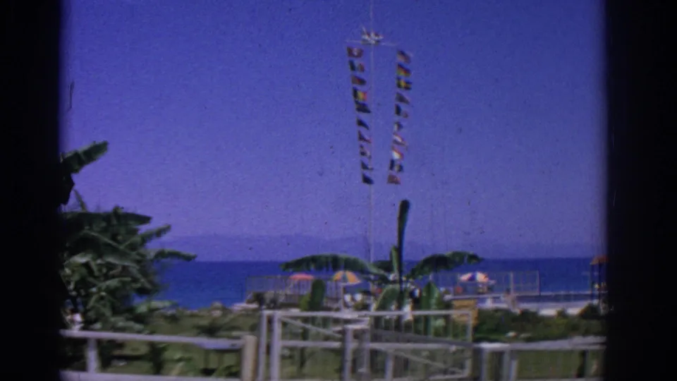 Rhodes 1962 waterfront frame showing tall striped flags above railings, umbrellas, plants, and the blue sea beyond.