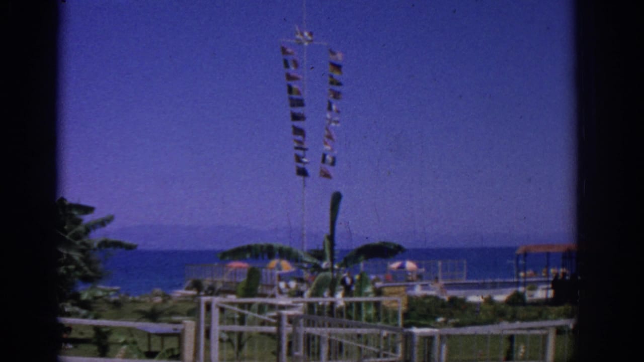 Seaside park seen through a dark window edge, with signal flags, palms, umbrellas, white railings, and blue sea.