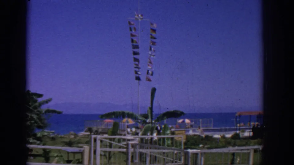 1962 Rhodes, Greece home-movie frame of a windy seaside park with tall signal flags above palms, umbrellas, railings, and the sea horizon.