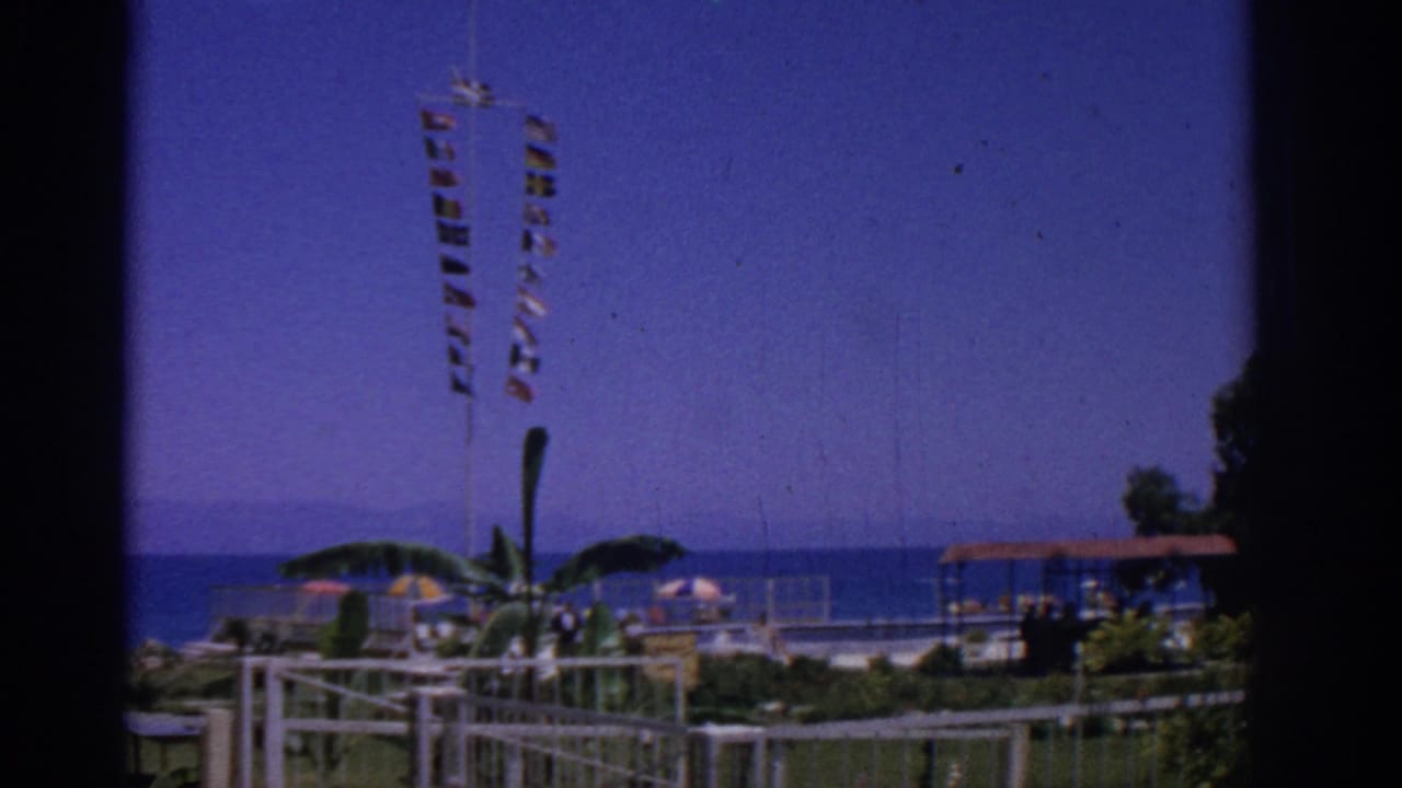 Windy beachfront park with signal flags, umbrellas, palms, railings, pavilion roof, and distant sea.