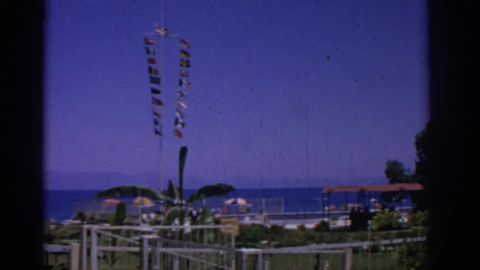 1962 Rhodes, Greece view across a beachfront park where signal flags flutter above umbrellas, palms, white railings, and a pavilion by the sea.
