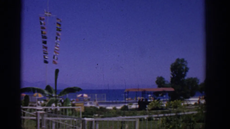 1962 Rhodes, Greece frame of a coastal park with signal flags at left, white railings, umbrellas, planted shrubs, a tree, pavilion, and blue water.