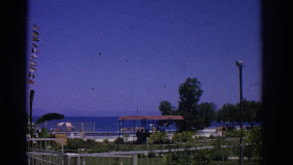 1962 Rhodes, Greece home-movie frame showing a seaside park with partial signal flags, umbrellas, pavilion roof, shrubs, a tree, lamp post, and open sea.