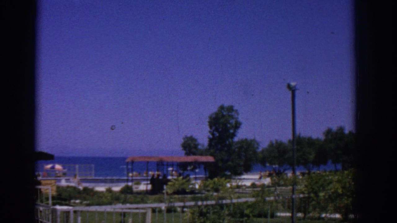 Seaside resort grounds seen through a dark window frame with blue water, trees, shelter, fence, and lamppost.