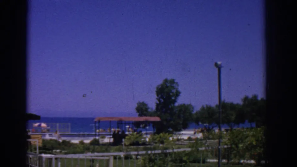 1962 Rhodes resort view through a window frame, showing blue sea, red-roofed shelter, trees, low fence, and a tall lamppost.
