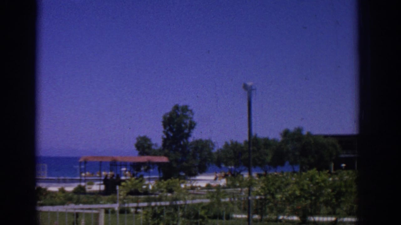 1962 Rhodes beachfront resort scene with blue ocean, a small red-roofed shelter, trees, lamppost, and distant people.