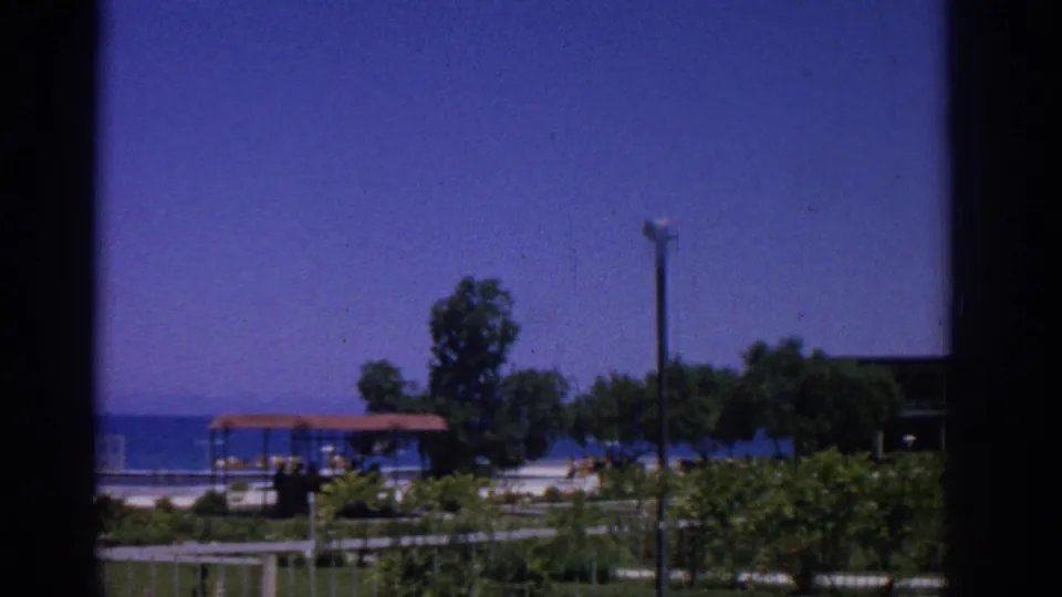 1962 Rhodes beachfront resort scene with blue ocean, a small red-roofed shelter, trees, lamppost, and distant people.