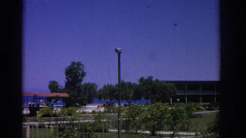 1962 Rhodes resort grounds with sea in the background, shelter at left, trees, low fence, lawn area, and a building edge.