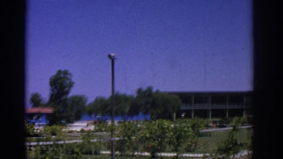 1962 Rhodes resort view showing a blue ocean horizon, trees, lamppost, fence, dense greenery, and part of a shaded building.