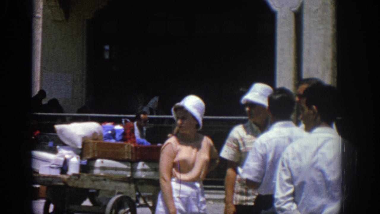 Adults in summer clothing stand near a loaded cart beneath shaded port arches.