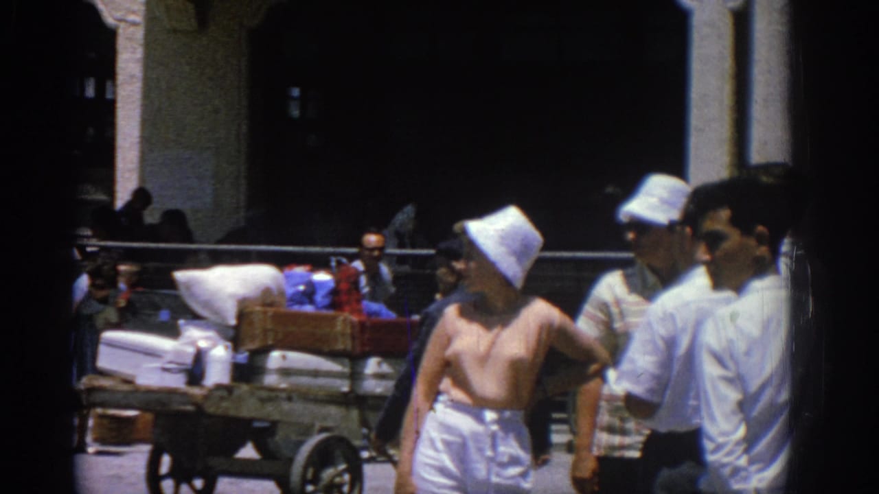 A woman in a white sunhat and nearby men stand by a loaded cart at the Rhodes port in 1962.
