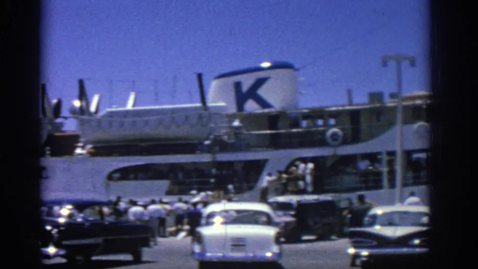 Classic 1960s cars and pedestrians stand near a docked passenger ship with a large K marking at Rhodes port.