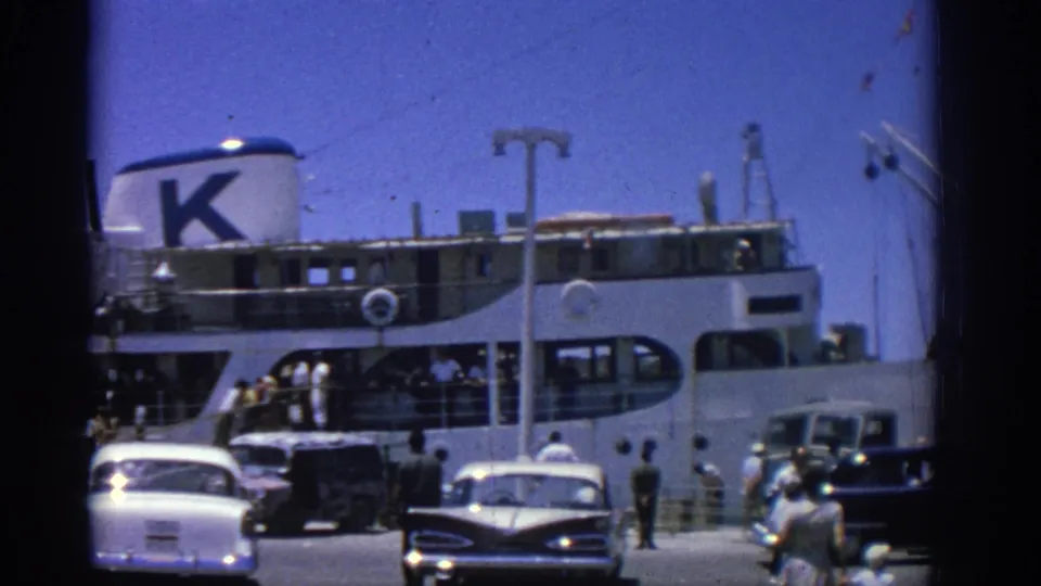 The Rhodes port scene shows a docked passenger vessel, period automobiles, and pedestrians moving along the waterfront in 1962.