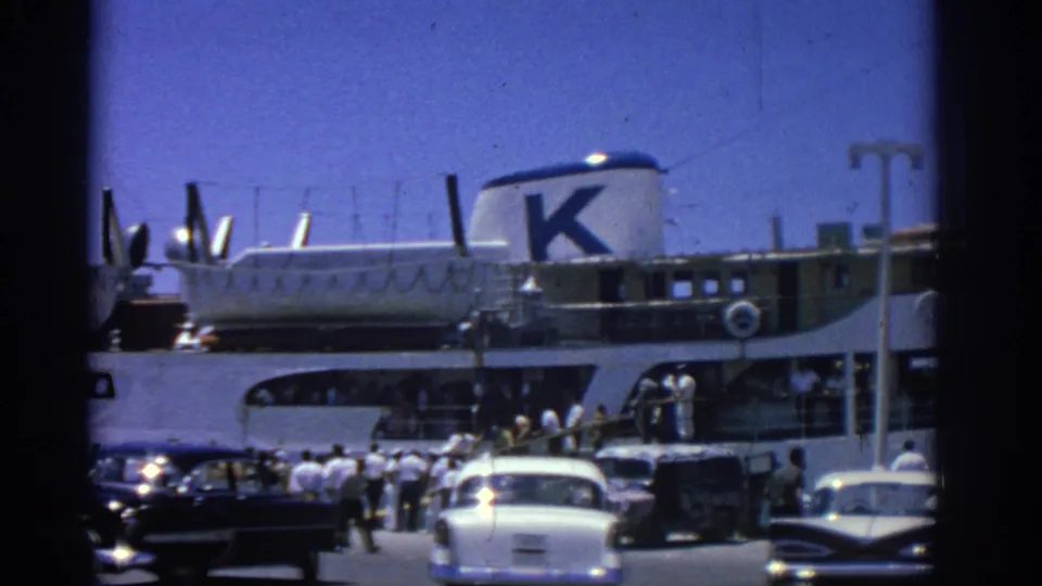 Rhodes, Greece, 1962 harbor view with passengers, early-1960s cars, and a white ferry marked with a large K at the dock.