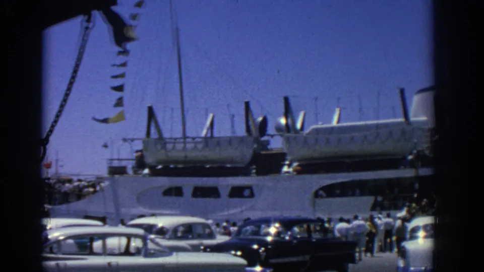 1962 Rhodes port activity with a ferry alongside the quay, flags above the frame, passengers gathered, and classic cars in front.