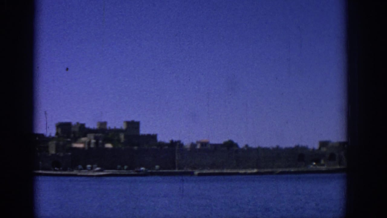 1962 Rhodes, Greece waterfront view with dark stone coastal buildings across calm blue water and visible 8mm frame edges.