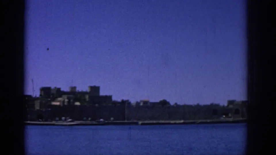 1962 Rhodes, Greece waterfront view with dark stone coastal buildings across calm blue water and visible 8mm frame edges.