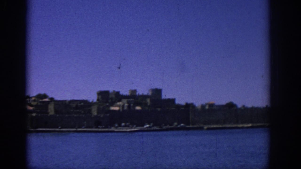 Rhodes waterfront in 1962, showing a low fortified coast, distant buildings, blue sea, and dark 8mm frame edges.