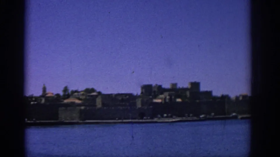1962 Rhodes harbor view with blue water in the foreground, a dark stone shoreline, and low buildings beneath open sky.