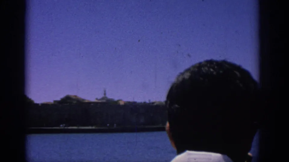 Final 1962 Rhodes waterfront frame with a person at lower right facing the sea and distant fortified shoreline.