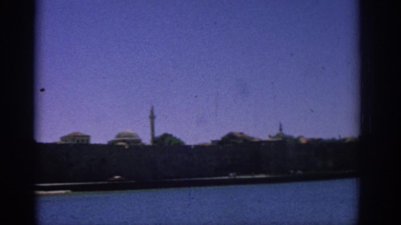 A steady 8mm view of the Rhodes waterfront shows domed buildings and a tall narrow tower across the harbor.