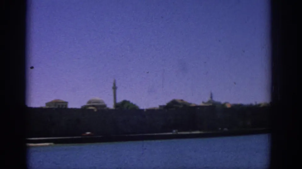 A steady 8mm view of the Rhodes waterfront shows domed buildings and a tall narrow tower across the harbor.