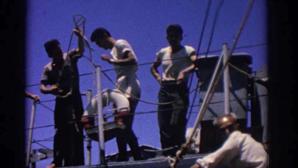 On a ship deck in Rhodes, several men work around ropes, railings, and deck equipment under a bright blue sky.