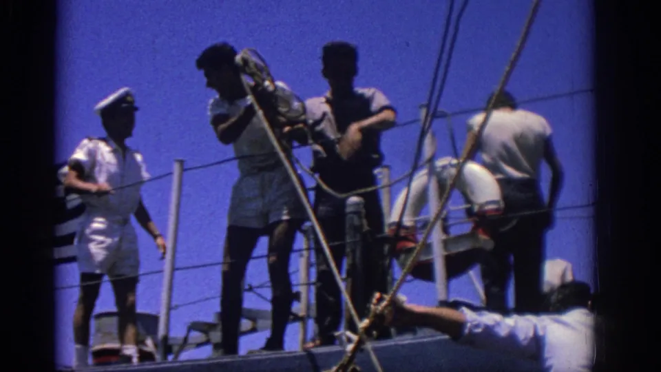 1962 Rhodes, Greece 8mm frame showing a uniformed sailor beside crew members, deck ropes, railing, and life preserver.