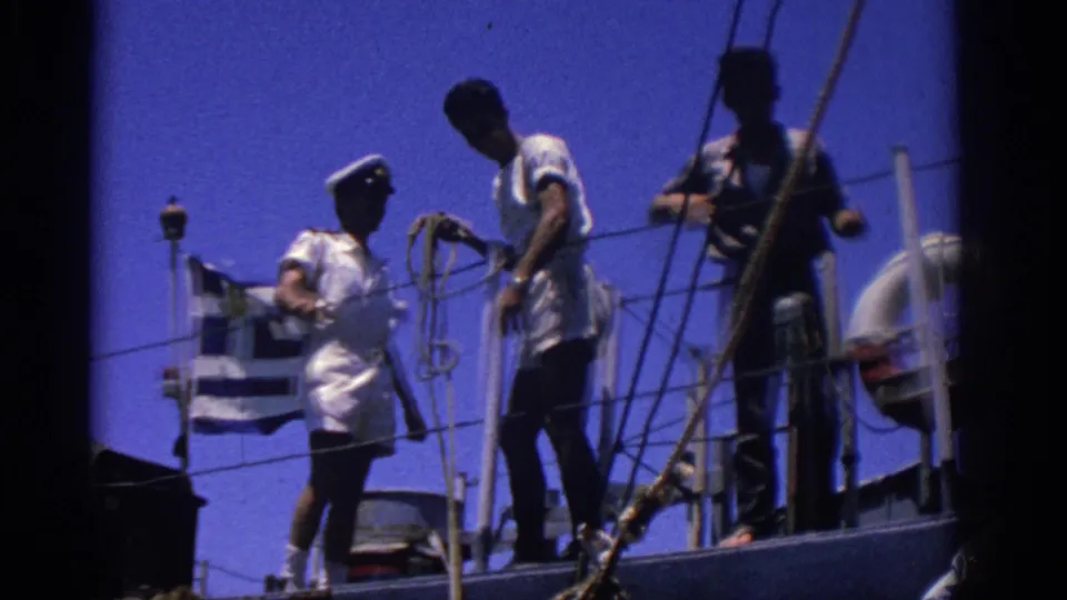 1962 Rhodes, Greece frame showing a Greek flag, sailors, rope rigging, railings, and a life preserver on deck.