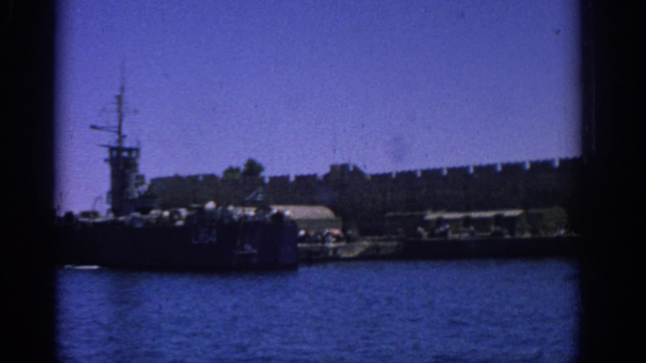 1962 Rhodes harbor view with a dark military vessel near a long stone fortress wall under blue sky.