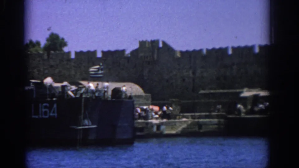 Closer 1962 Rhodes harbor frame showing the L164-marked vessel, people on the dock, and the fortress wall behind.