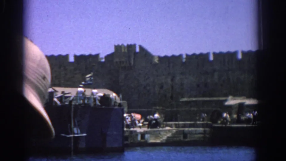 1962 Rhodes harbor frame with the L164 vessel, visible people, dock activity, stone wall, and a blurred foreground obstruction.
