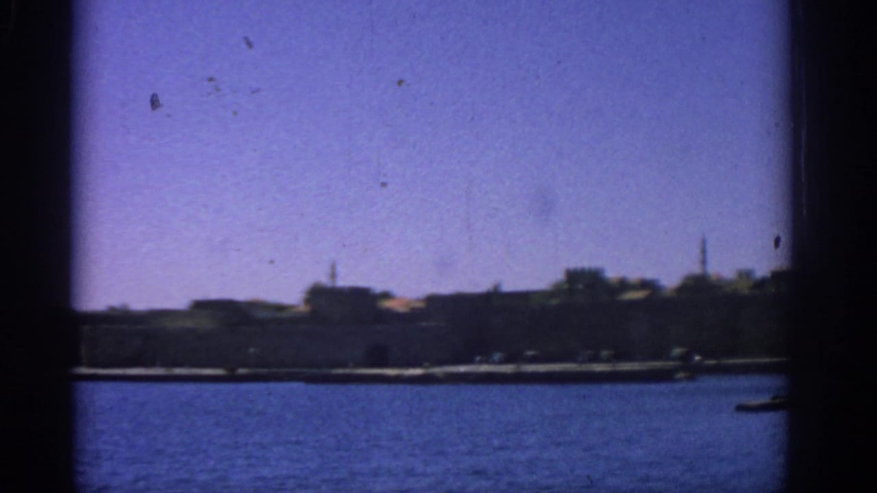 1962 Rhodes harbor view from a boat, showing blue water, distant stone walls, low buildings, and tower silhouettes.