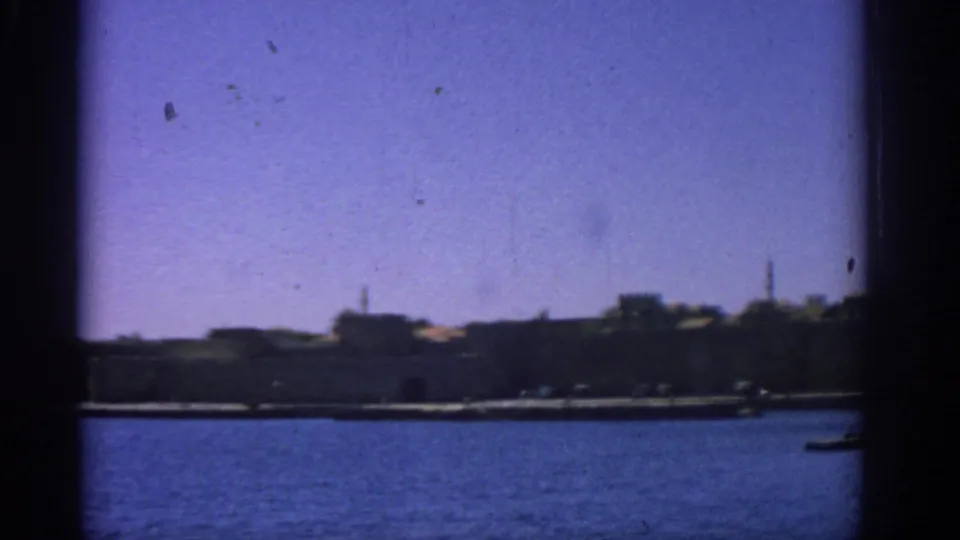 1962 Rhodes harbor view from a boat, showing blue water, distant stone walls, low buildings, and tower silhouettes.