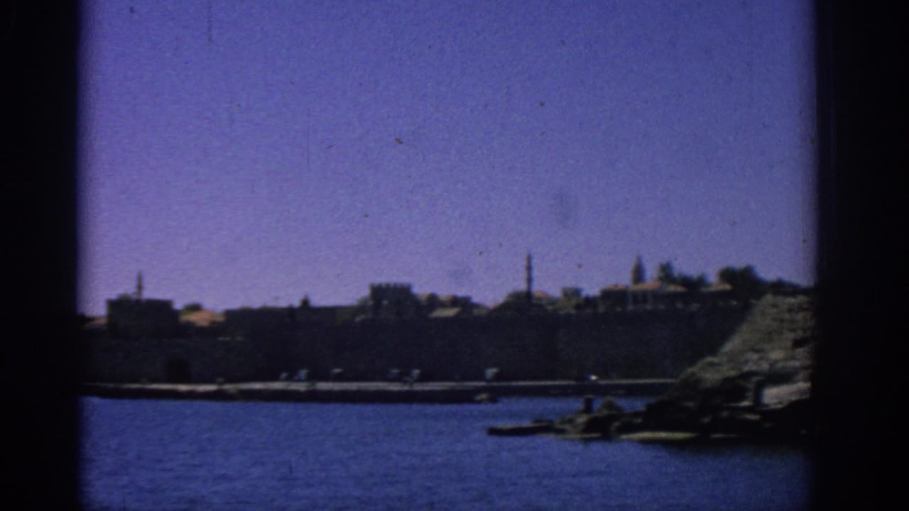 Rhodes coastal harbor from a moving boat, with stone waterfront walls, small boats, and rocky shoreline at the right edge.