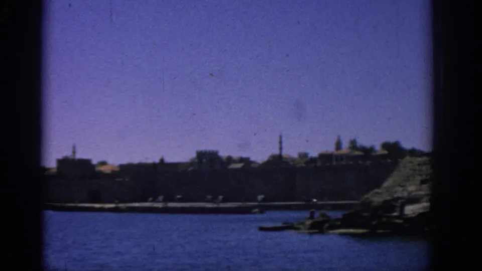 Rhodes coastal harbor from a moving boat, with stone waterfront walls, small boats, and rocky shoreline at the right edge.