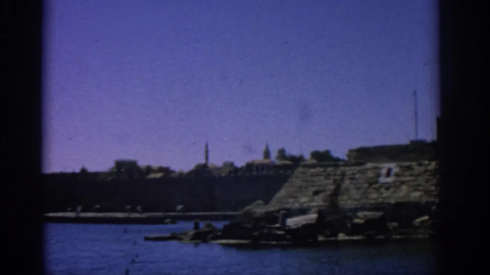 1962 Rhodes waterfront seen from the water, with a long stone wall, small boats, distant towers, and sunlit coastal structures.