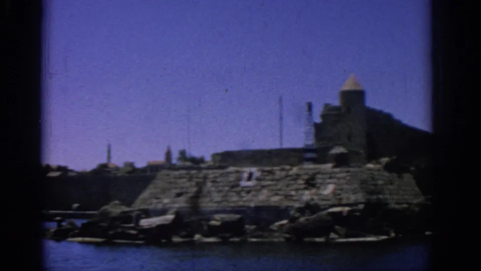 Rhodes harbor view from 1962 showing blue water, stone fortifications, rocky shore, and a towered building at the right.