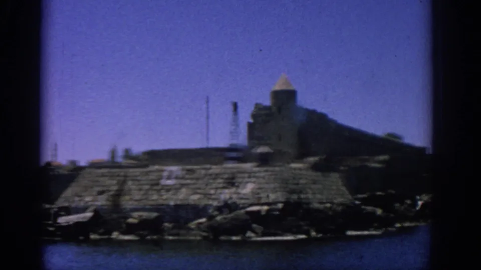1962 8mm view of a Rhodes coastal fortress with a tower, rocky edge, sea and clear blue sky.