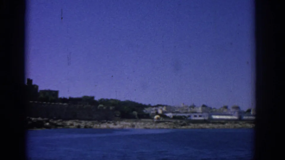 1962 Rhodes waterfront view with low fortification, distant buildings, sea and broad blue sky.