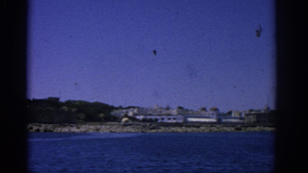 Coastal buildings and rocky shoreline seen across blue water through a dark film-frame border.