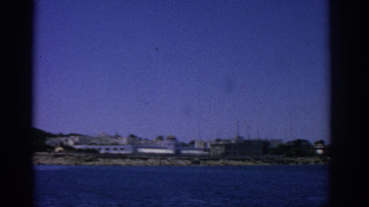Distant coastal town with pale buildings and a rocky edge beyond blue water.