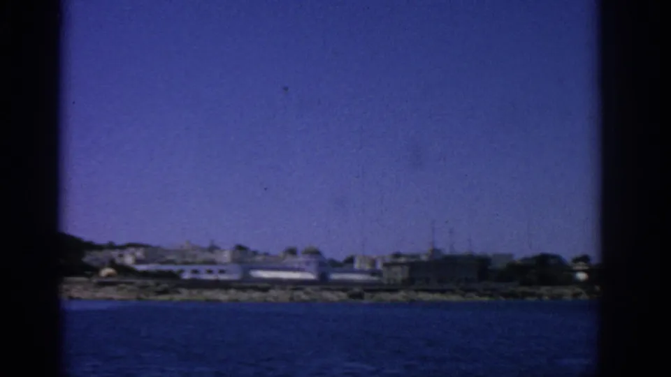 Rhodes coastline in 1962, with pale waterfront structures and a rocky shore viewed from a boat on blue water.