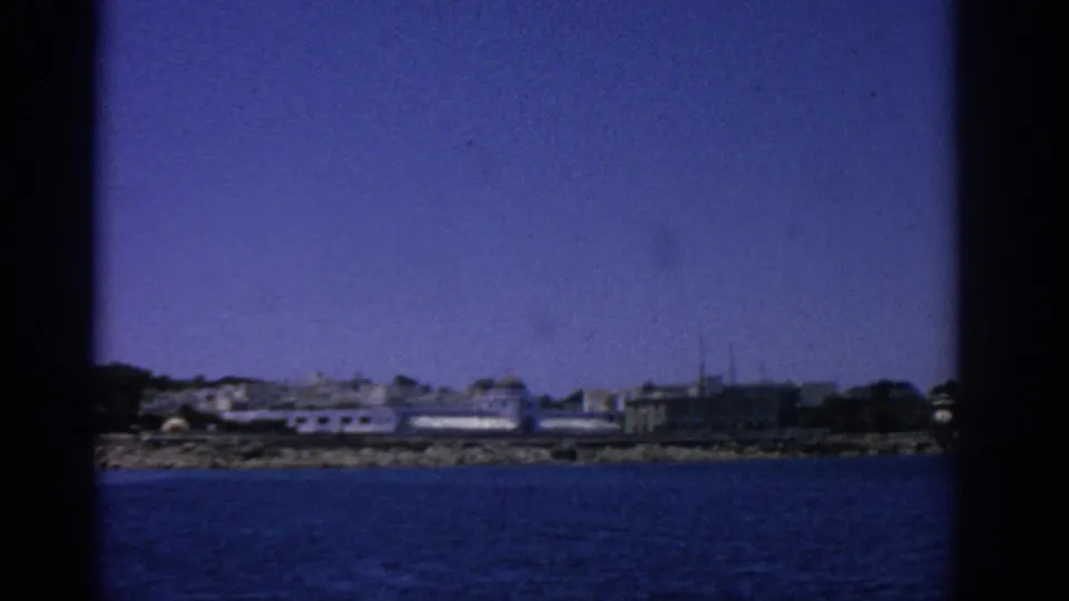 Wide 1962 Rhodes shoreline view from the water, showing low coastal buildings, rocks, and a strong blue film cast.