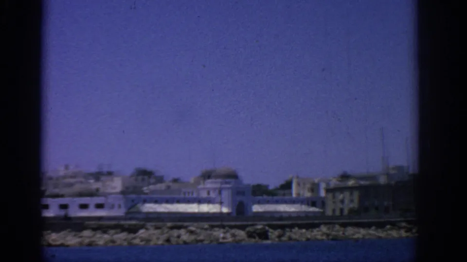Closer 1962 8mm view of Rhodes waterfront buildings, rocky shoreline, and blue water with dark frame edges.