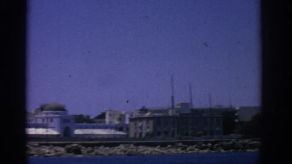 1962 Rhodes home-movie frame showing pale waterfront buildings and a rocky coast seen from the water.