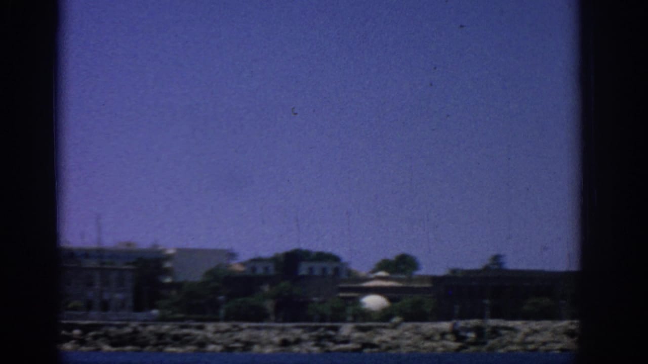 Rocky coast with low buildings and trees seen across the water.