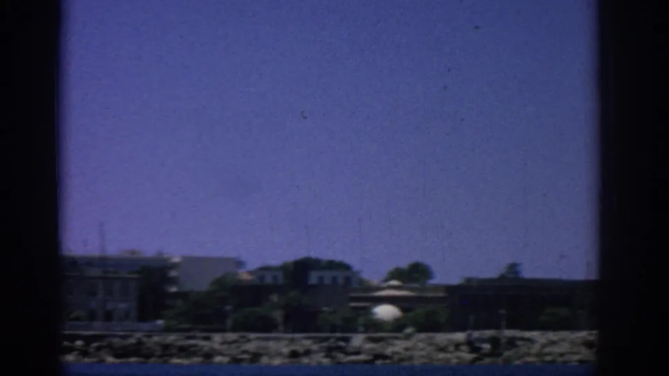 1962 Rhodes shoreline view from a boat, with low coastal buildings, trees, and open sky.
