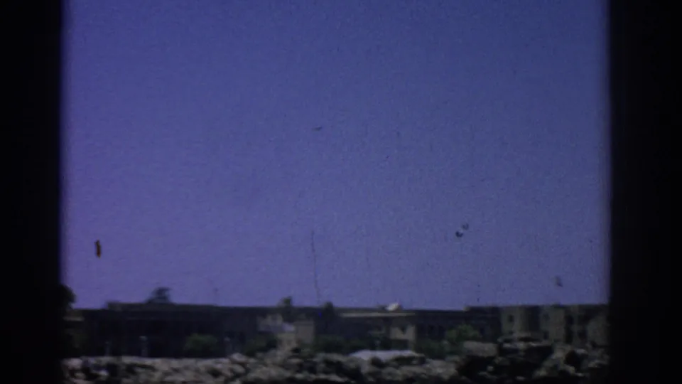 1962 Rhodes coastal frame with distant waterfront structures, rocky foreground, and expansive sky.